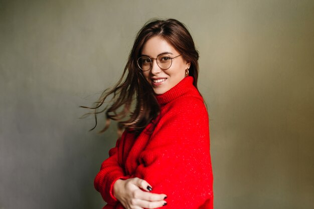 Snapshot of good-natured, tanned girl on gray wall. European model in red sweater smiles friendly.