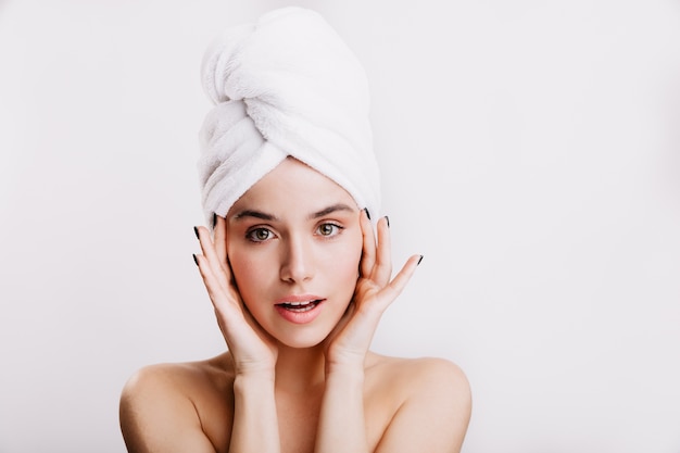 Snapshot of healthy beautiful girl in white towel on her head. Woman with green eyes touches her face.