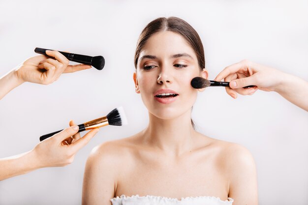 Snapshot of model with healthy skin. Many hands with makeup brushes reaching out to face of young woman.