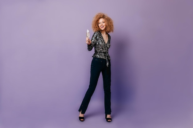 Бесплатное фото Снимок стройной длинноногой девушки в темных джинсах, блестящей блузке, позирующей с бокалом шампанского на фиолетовом фоне