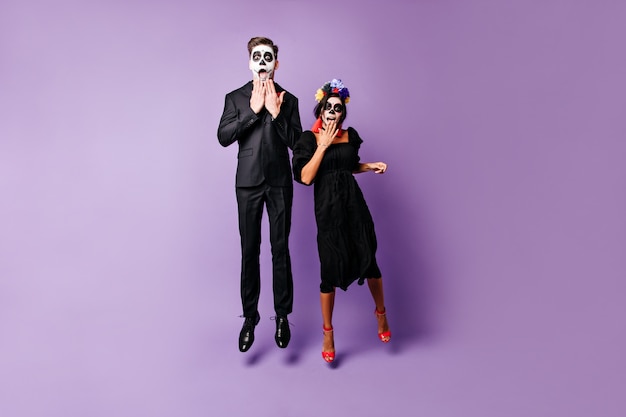 Free photo snapshot of young slender guy and girl with painted faces in black outfits. couple from mexico looking surprised into camera, jumping on isolated background.