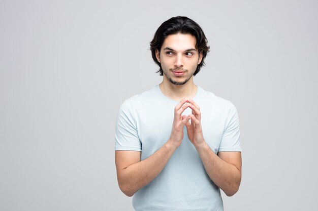 Sneaky young handsome man keeping hands together looking at side planning something evil isolated on white background with copy space