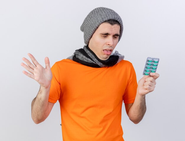 Sneezing young ill man wearing winter hat with scarf holding pills and spread hand isolated on white