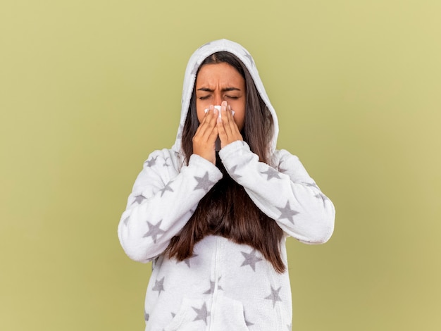 Free photo sneezy young ill girl putting on hood wiping nose with napkin isolated on olive green