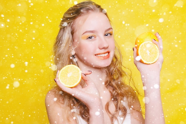 눈, 겨울, 크리스마스, 감정, 건강, 사람, 음식, 미용 개념 - 뷰티 모델 소녀는 쥬시 오렌지를 먹습니다. 주근깨가 있는 아름다운 즐거운 십대 소녀. 노란색 메이크업. 눈 배경 위에 프리미엄 사진