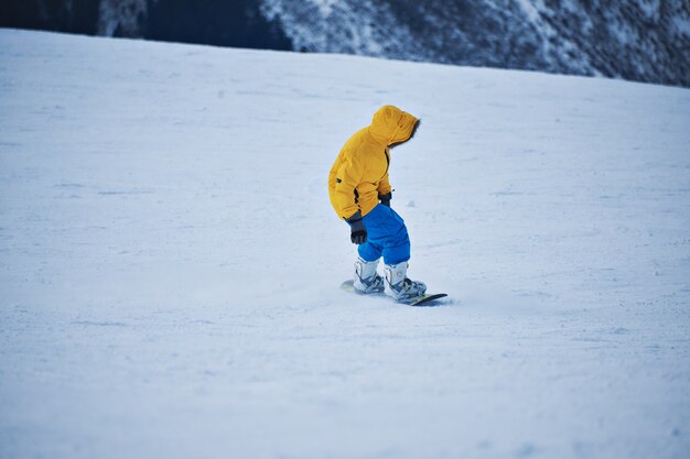 Сноубордист в ярко-желтой куртке и синих штанах смотрит вниз на снежный склон перед началом катания в солнечный зимний день на горнолыжном курорте