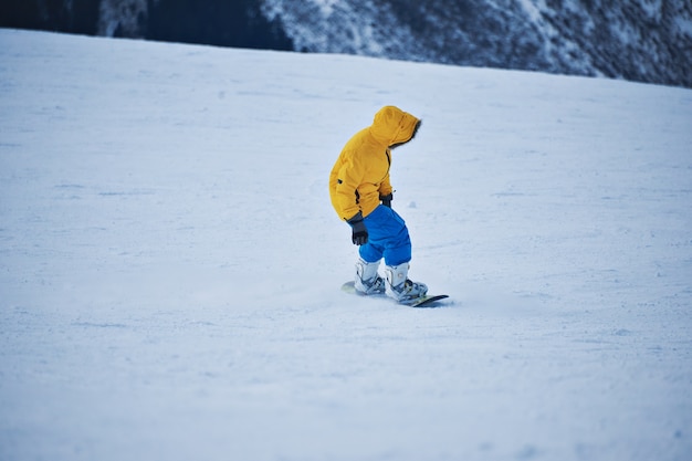 Snowboarder in bright yellow parka and blue pants looks down on snow slope before start to ride at sunny winer day in mountain ski resort