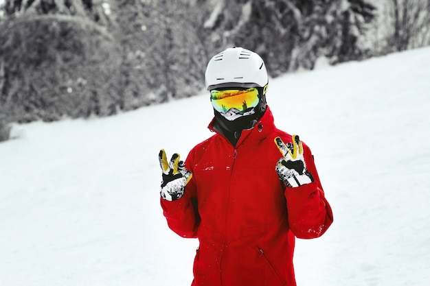 Foto gratuita snowboarder in giacca rossa, casco bianco e occhiali gialli posa sulla collina