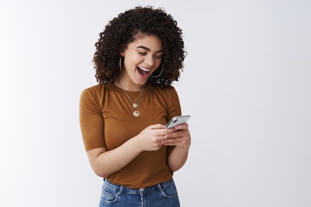 So awesome simple. Cheerful attractive young relaxed curly dark hair stylish girl laughing open mouth excited reading daring message hold smartphone reacting amused funny video online look display