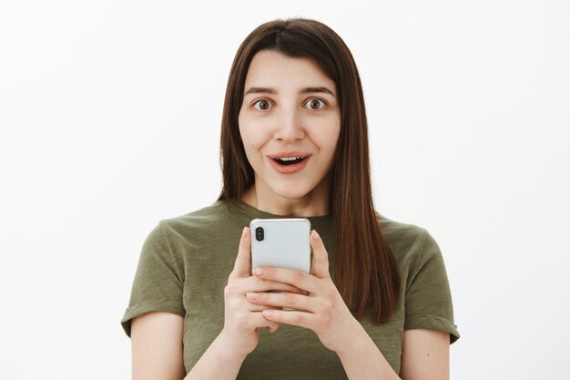 So cool best phone ever. Portrait of impressed and fascinated happy speechless brunette in 20s holding smartphone and smiling astonished as excited of cellphone features over gray wall