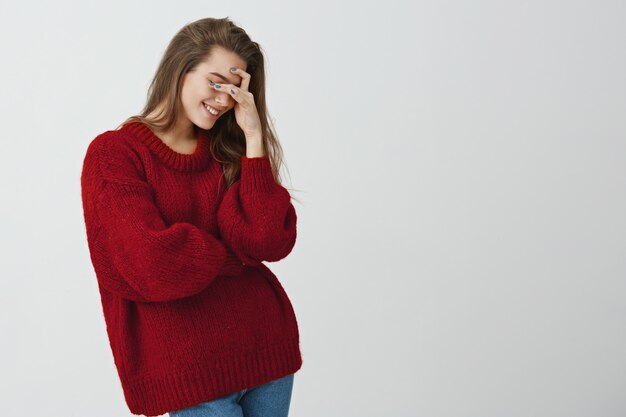 So dumb but so sweet. Studio shot of attractive stylish woman in red loose sweater making facepalm, holding hand on eyes and smiling, coming to terms with stupid sense of humour of boyfriend