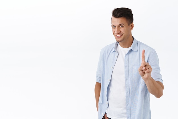 Not so fast, one more thing. Portrait of handsome charismatic man, shaking index finger and smiling, rejecting or prohibit something, have one idea, pointing up, standing white wall