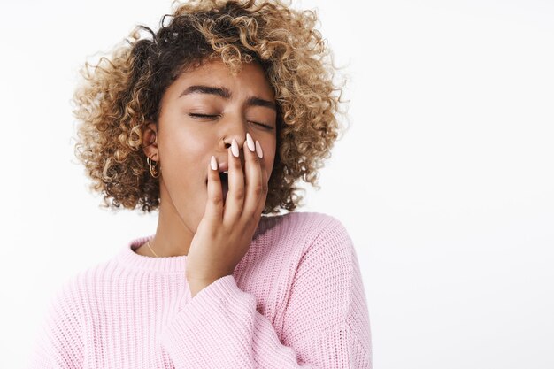 So sleepy. Portrait of cute and tender african-american woman with curly blond haircut in pink sweater yawning and cover opened mouth with palm cute, close eyes feeling tired after cool awesome party