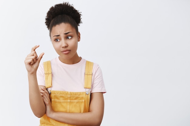 So small that is shame. Portrait of displeased upset and unimpressed African-American female student in yellow trendy overalls, frowning while gazing at fingers, shaping something little and tiny