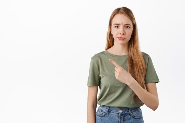 Not so sure Skeptical young woman smirk and frown from doubts pointing left at something average judging bad promo deal standing over white background