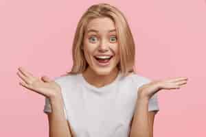 Free photo so, what? happy woman shrugs shoulders, looks joyfully with her blue eyes, dressed casually, isolated over pink wall. hesitant cheerful blonde female expresses positive emotions and feelings