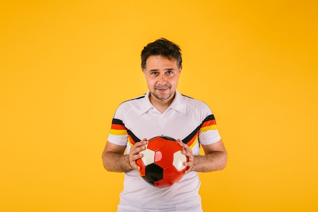 Футбольный фанат, одетый в белую футболку с черными, красными и желтыми полосами, держит мяч в руках Premium Фотографии