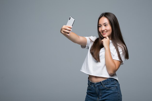 Selfieを取るか、灰色の背景に携帯電話を使用してビデオ通話で話す社交的な美しいアジアの少女
