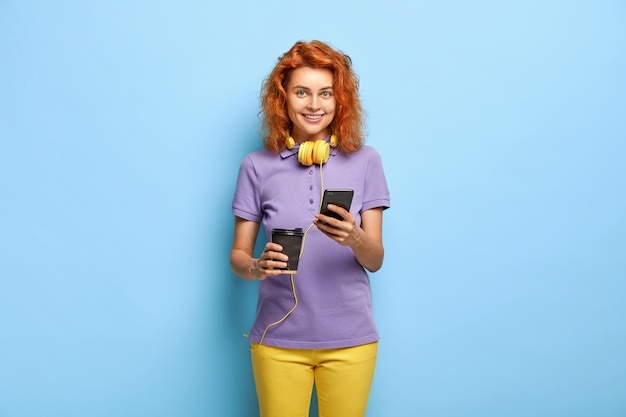Sociable communicative girl sends text messages via smart phone, drinks coffee from paper cup