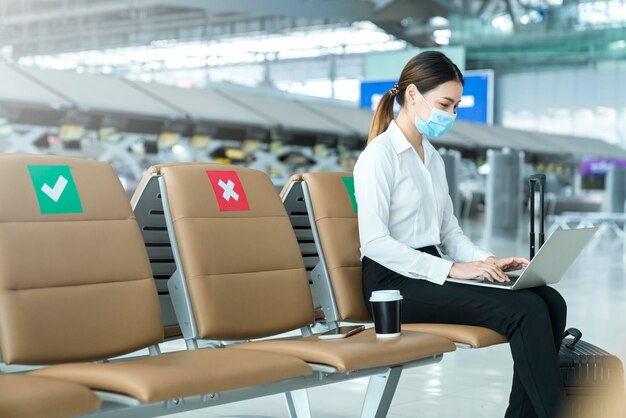 Social distancing businesswoman wearing face mask sit working with laptop keeping distance away from each other to avoid covid19 infection during pandemic Empty chair seat red cross shows new normal