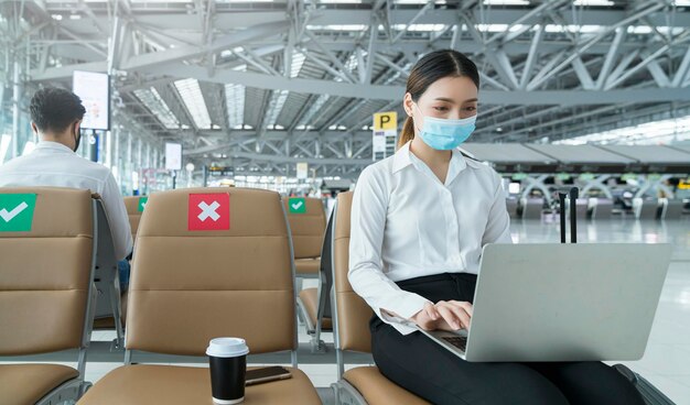 Social distancing businesswoman wearing face mask sit working with laptop keeping distance away from each other to avoid covid19 infection during pandemic Empty chair seat red cross shows new normal