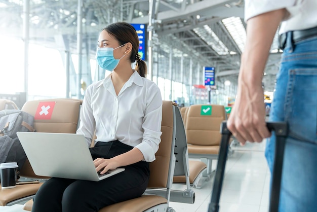 Social distancing businesswoman wearing face mask sit working with laptop keeping distance away from each other to avoid covid19 infection during pandemic Empty chair seat red cross shows new normal
