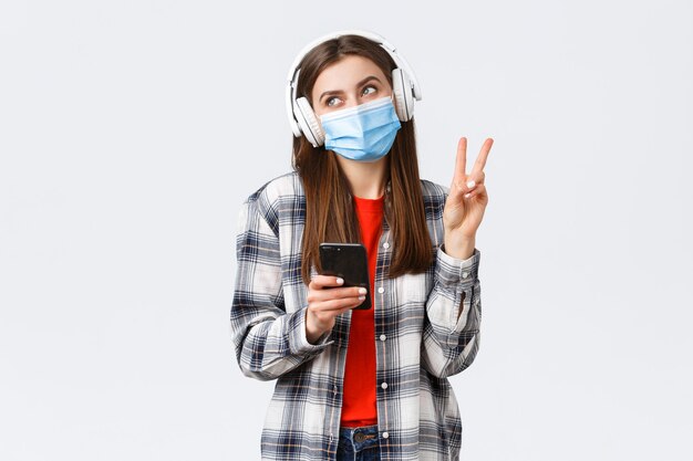 Social distancing, leisure and lifestyle on covid-19 outbreak, coronavirus concept. Happy cheerful silly girl in medical mask and headphones, show peace sign smiling, holding mobile phone.