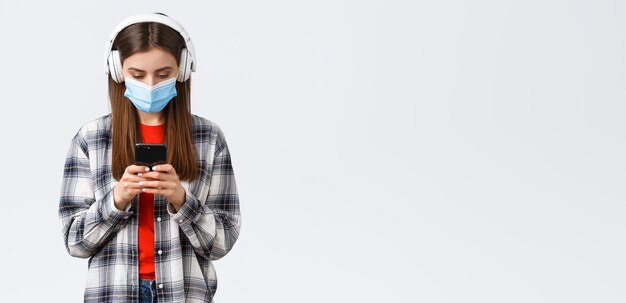 Foto gratuita tempo libero e stile di vita di distanza sociale sul concetto di coronavirus dell'epidemia di covid19 ragazza adolescente carina studentessa in cuffia che seleziona una canzone nel telefono cellulare scarica l'app musicale sfondo bianco