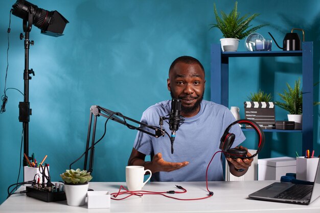 Social media influencer presenting professional gaming headset to audience sitting at desk with laptop and video recording setup. Vlogger reviewing a pair of audio headphones explaining features.