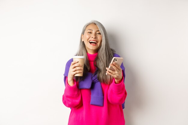 Социальная сеть. Счастливая азиатская старшая женщина пьет кофе и держит смартфон, смеясь над камерой, стоя на белом фоне