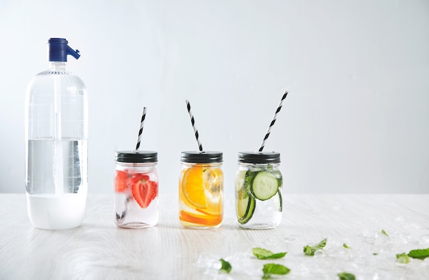 Soda syphon bottle near three rustic jars with icy fresh beverage from strawberry, orange, lime, mint, cucumber and sparkling water