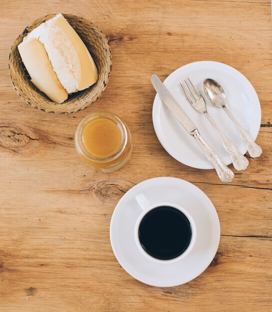 Soft bread in wicker basket; jam; coffee cup and cutlery set on white plate against wooden backdrop