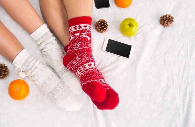 Soft photo of woman and man on the bed with phone and fruits, top view point. Female and male legs of couple in warm woolen socks.