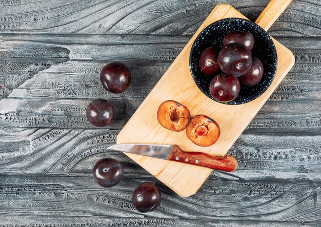 Foto gratuita prugne molli con un coltello da frutta in un piatto e un bordo di legno su fondo di legno grigio, disposizione piana.