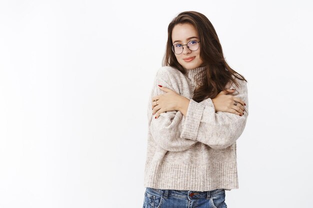 Soft and tender happy young sensual woman in glasses and sweater hugging herself as warming up in cozy outfit