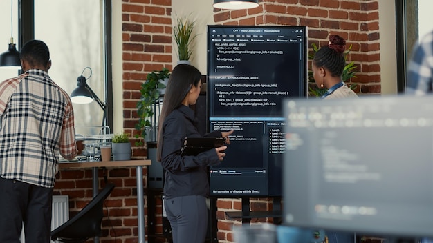 Инженер-программист держит цифровой планшет, анализируя код на настенном экране телевизора, объясняя ошибки коллеге-программисту в стартовом офисе. Команда кодеров, сотрудничающих над проектом инновационной группы ИИ.