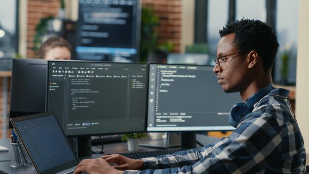 Sofware developer thinking while touching beard while typing on laptop sitting at desk with multiple screens parsing code. Focused database admin working with team coding in the background.