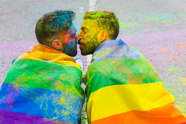 Soiling in paint gay pair kissing wrapped in rainbow flag on LGBT pride parade