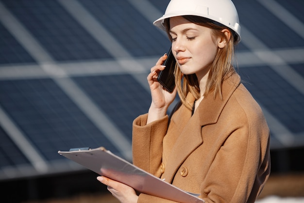 太陽光発電所。工場で働く若い女性エンジニア。彼女は電話で話し、仕事をしています。書類を持ったヘルメットをかぶった女性。