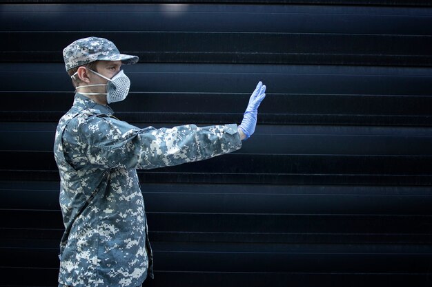 Солдат в камуфляжной форме в защитных перчатках и маске показывает знак остановки рукой на черном фоне