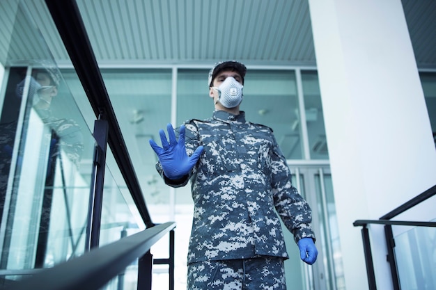 ゴム手袋と顔保護マスクを備えた軍服を着た兵士が病院を守り、一時停止の標識を身振りで示す