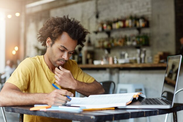 Торжественный темнокожий афроамериканский студент на своем рабочем месте смотрит в тетрадь и пишет заметки, готовясь к выпускным экзаменам в университете. Сосредоточенный красивый парень, работающий в кафе во время перерыва