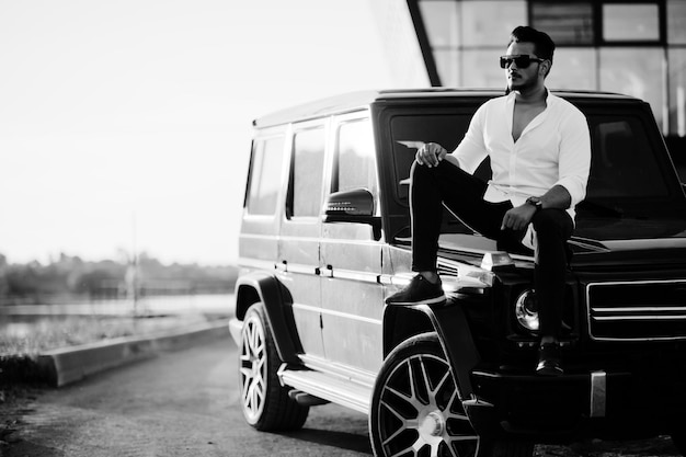 Solid asian man in white shirt and sunglasses posed near black mafia suv car