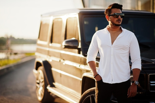 Solid asian man in white shirt and sunglasses posed near black mafia suv car