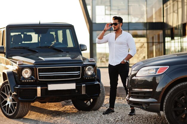 Solid asian man in white shirt and sunglasses posed near black mafia suv cars