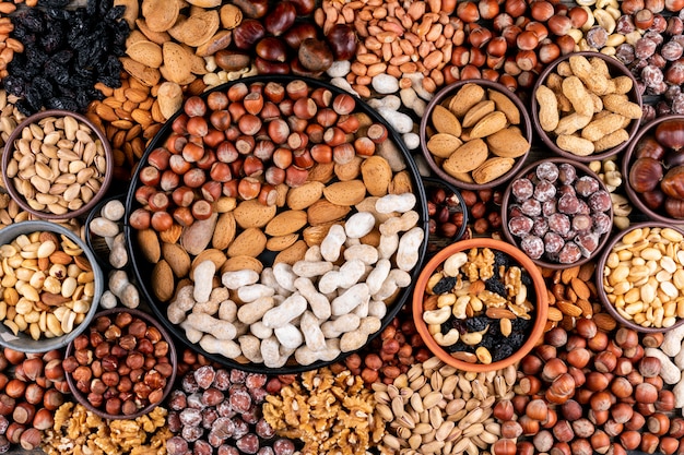 Some of assorted nuts and dried fruits with pecan, pistachios, almond, peanut, cashew, pine nuts in a different bowls and black pan flat lay.