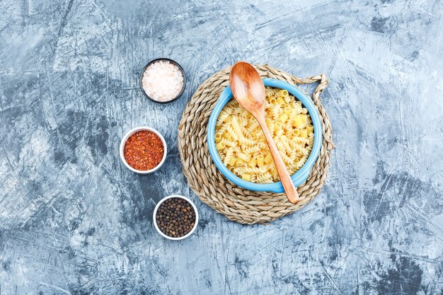 Free photo some assorted pasta with spices, wooden spoon in a bowl on grey plaster and wicker placemat background, top view.