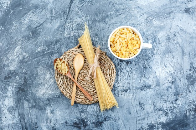 Some assorted pasta with wooden spoons in bowl on grey plaster and wicker placemat background, top view.