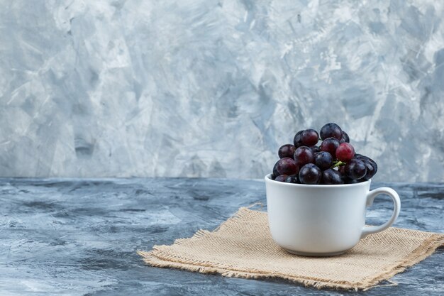 Some black grapes in a white cup on grunge and piece of sack background, side view.