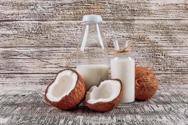 Some bottles of milk with divided in half coconut on white wooden background, side view.